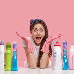 Horizontal shot of amazed overjoyed woman spreads hands, wears protective gloves, sits at table with many detergents, advertises good cleaning supplies for tidying up, isolated over pink background