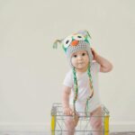 A cute baby wearing an owl hat standing in a basket, smiling and playful.